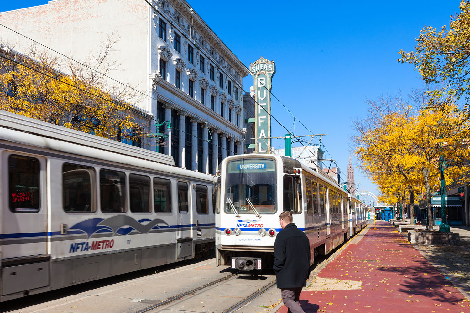 Man crosses street while two Metro Rail trains pass in front of Sheas Performing Arts Center in downtown Buffalo.